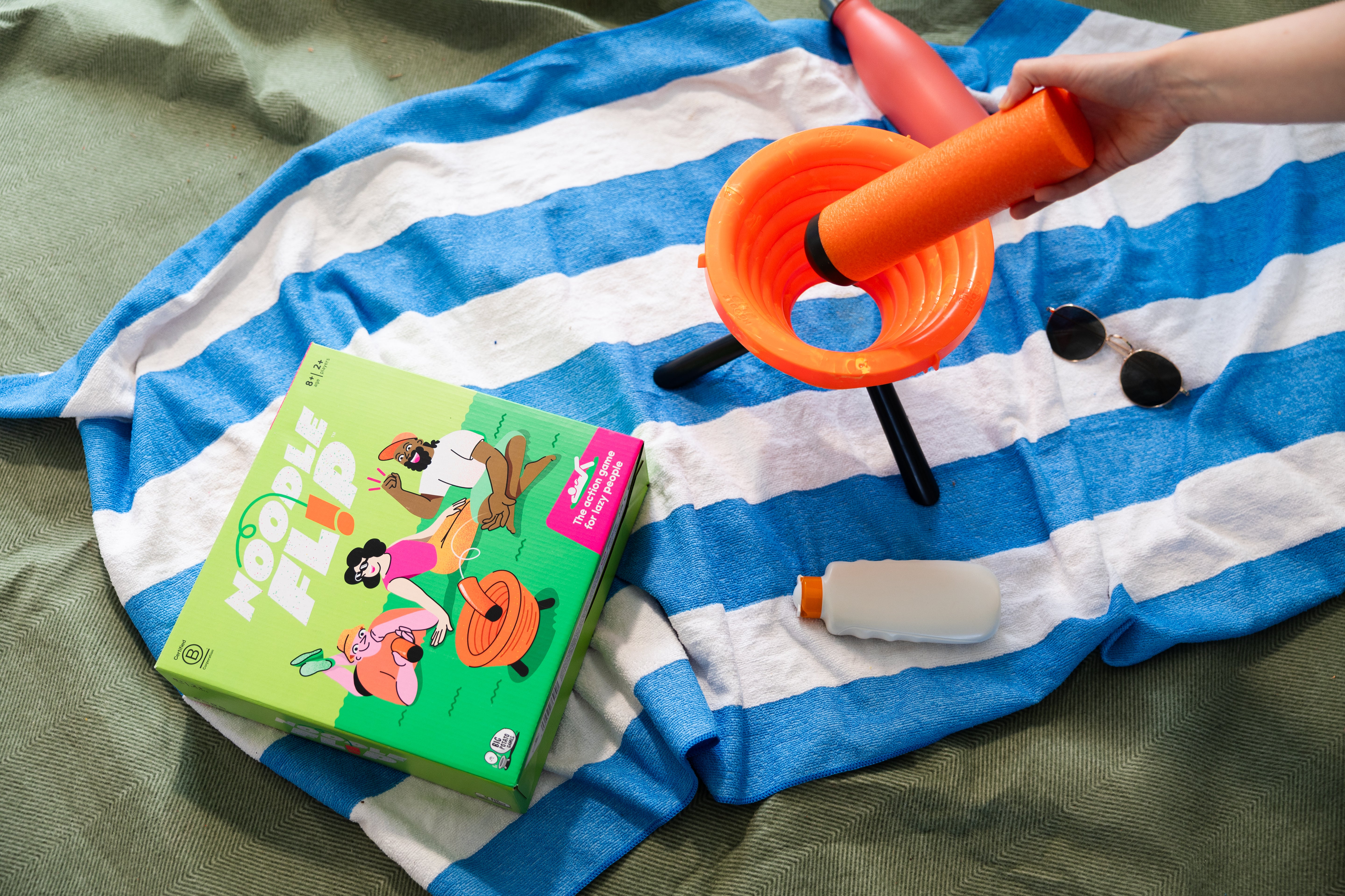 Big Potato Games Noodle Flip box on a beach towel with a hand holding the noodle in frame.