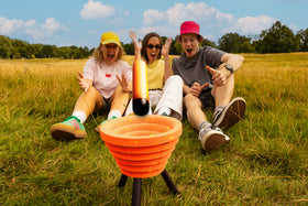 Three people react excitedly while playing Noodle Flip in a grassy field, as an orange foam noodle lands in the target bowl.