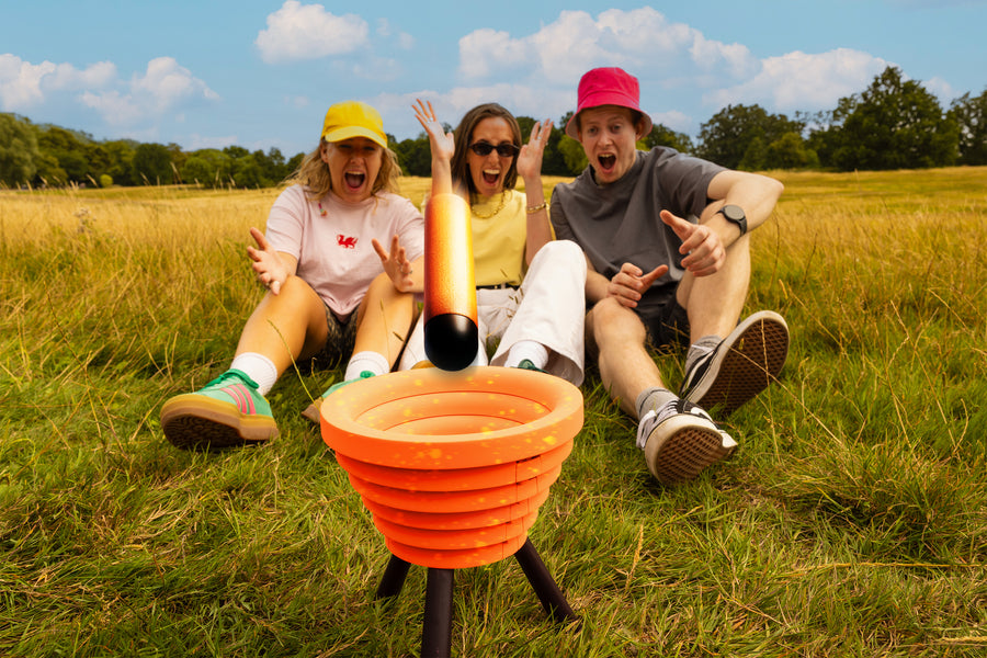 Three people react excitedly while playing Noodle Flip in a grassy field, as an orange foam noodle lands in the target bowl.