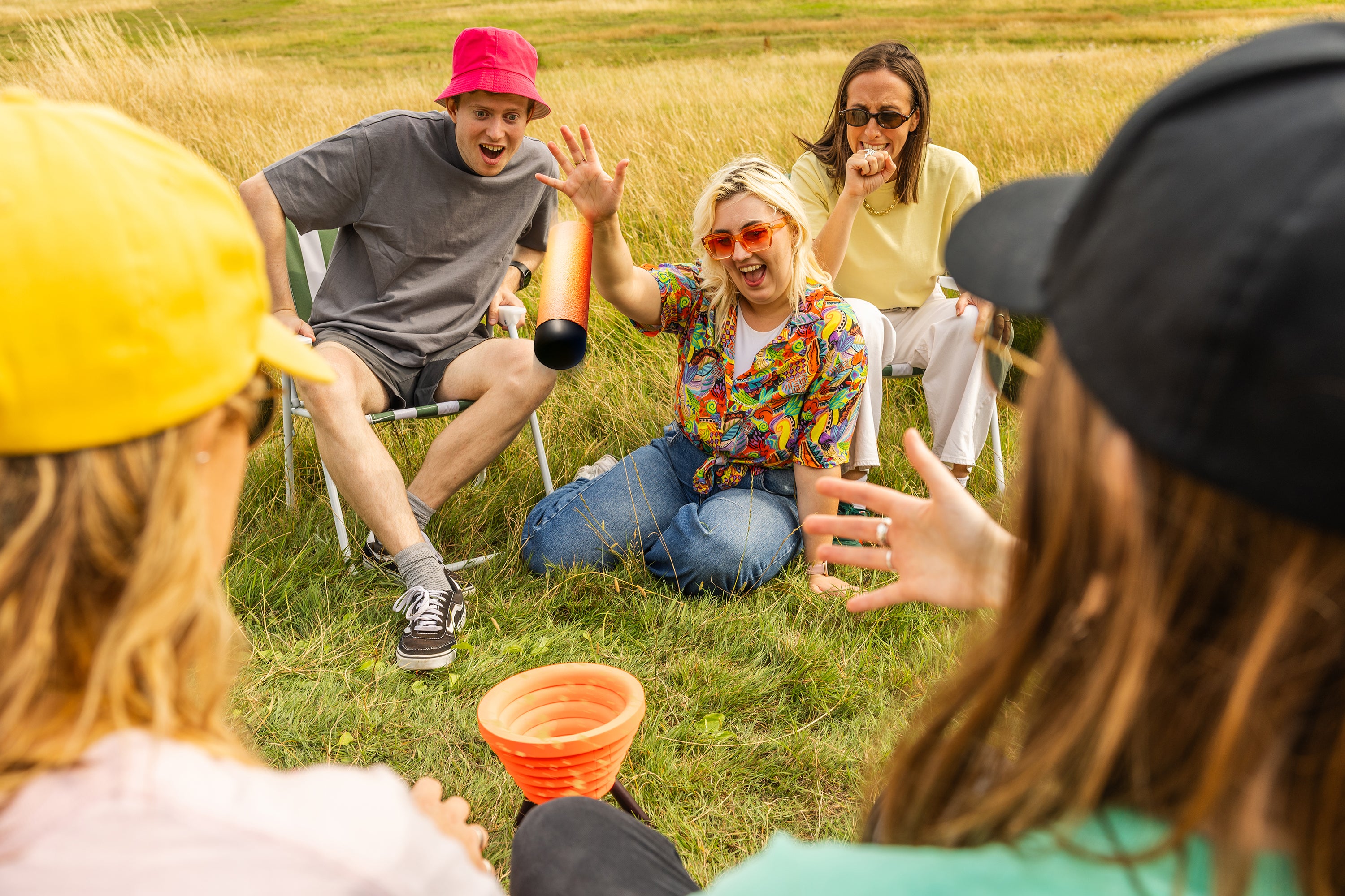 A group of friends play Noodle Flip outdoors in a grassy field, reacting as an orange foam noodle is flipped toward the target bowl in the middle of the group.