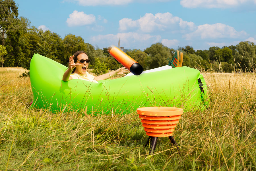A person lounges on a bright green inflatable chair in a grassy field while playing Noodle Flip, reacting as an orange foam noodle flies toward the target bowl.