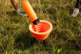 An orange foam noodle is flipped toward the target bowl in Noodle Flip, set up outdoors in long grass with players standing nearby.