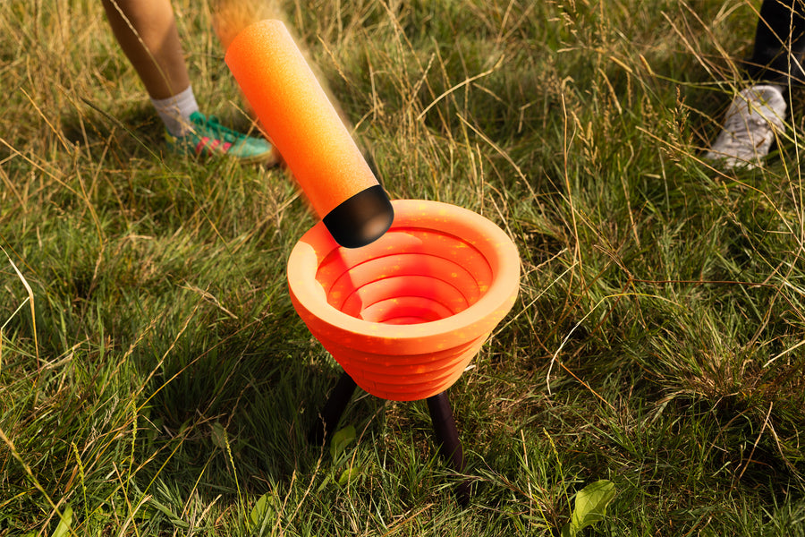 An orange foam noodle is flipped toward the target bowl in Noodle Flip, set up outdoors in long grass with players standing nearby.