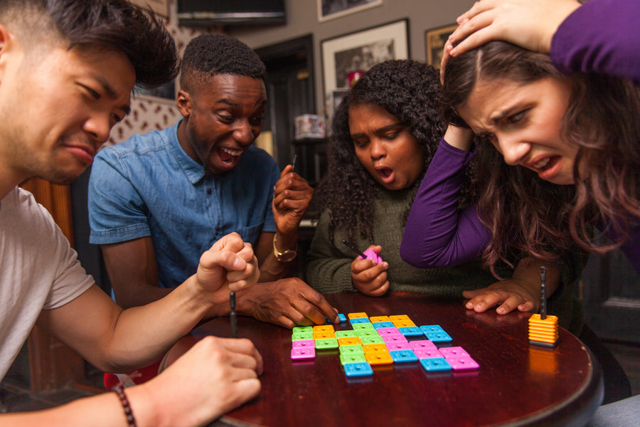 Four people gathered around a table playing a colorful board game in a dimly lit indoor setting. The game features bright pink, green, yellow, and blue tiles arranged in a grid on a dark wooden surface.